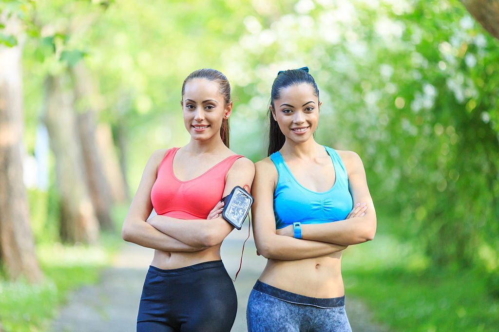 Two sportswomen posing arms crossed in park. 